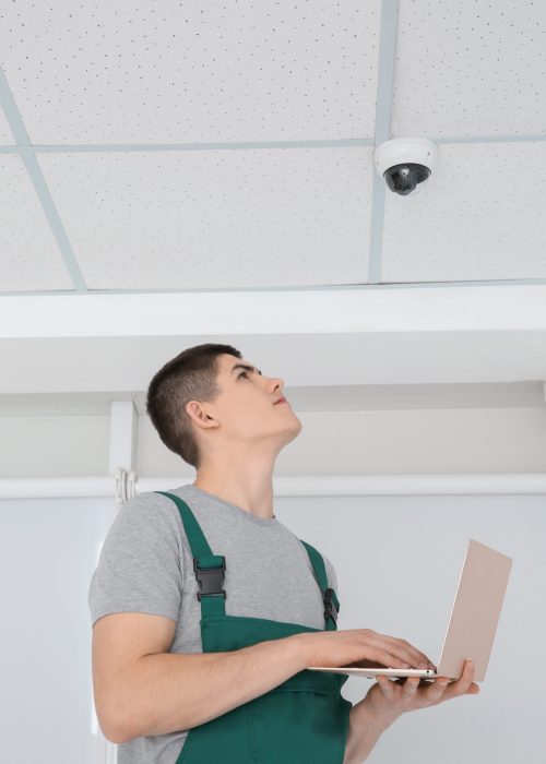 Technician with laptop installing CCTV camera on ceiling indoors, low angle view